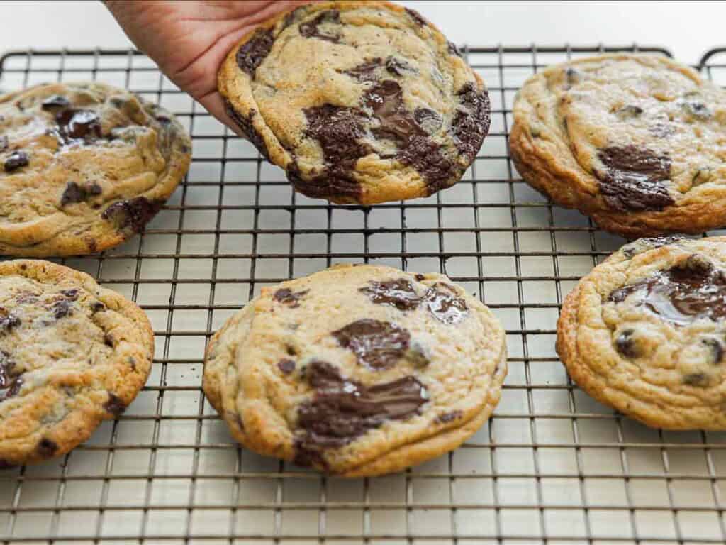 A hand lifts a freshly baked vegan chocolate chip cookie from a cooling rack with five other cookies, each showing melted chocolate chunks and a golden-brown surface.